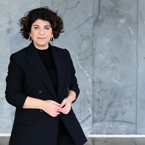 Eine Frau mit schwarzen Haaren steht vor einer grauen Wand.